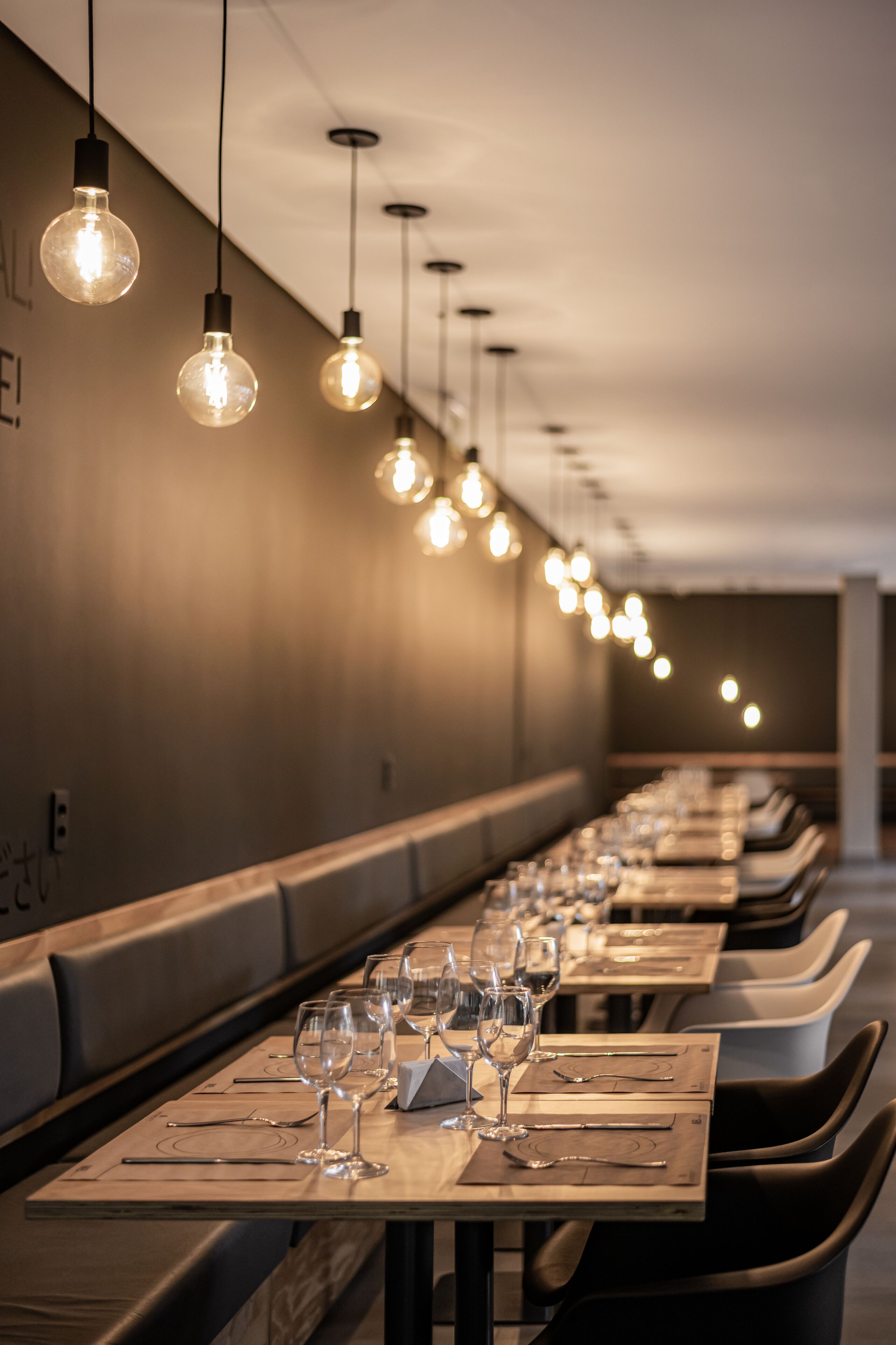 A long dining table with multiple chairs, each set with cutlery and wine glasses, in a dimly lit room with hanging light bulbs.