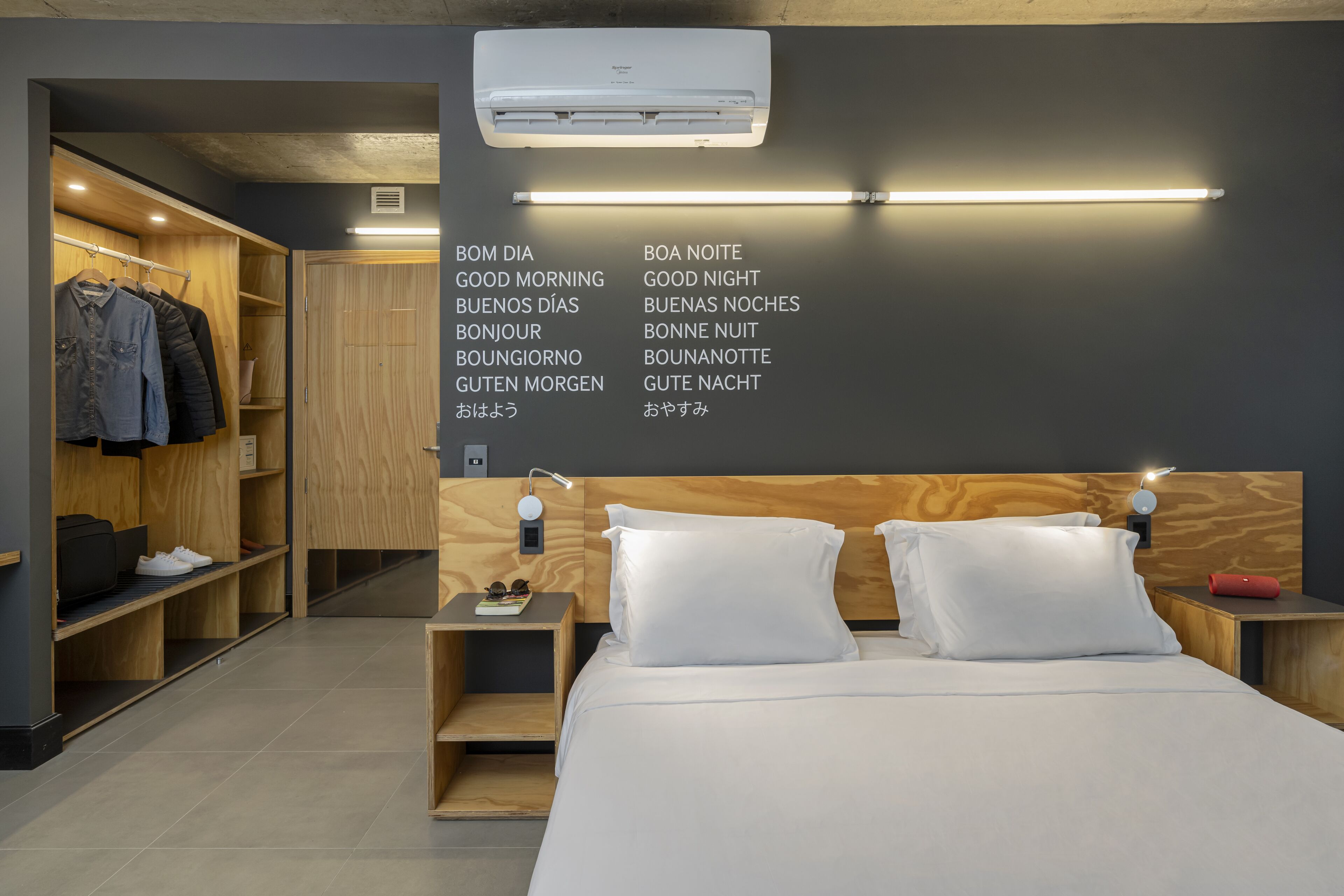 Una habitació d'hotel moderna amb un capçal de llit de fusta, un llit amb roba de llit blanca, una nit de nit amb una làmpada, un armari amb roba i una paret amb cartells multilingües de bon dia i bona nit.