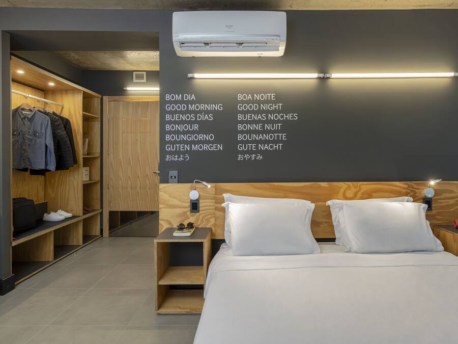 Una habitación de hotel moderna con cabecera de madera, cama con ropa blanca, mesita de noche con lámpara, armario con ropa y pared con letreros bilingües de buenos días y buenas noches.