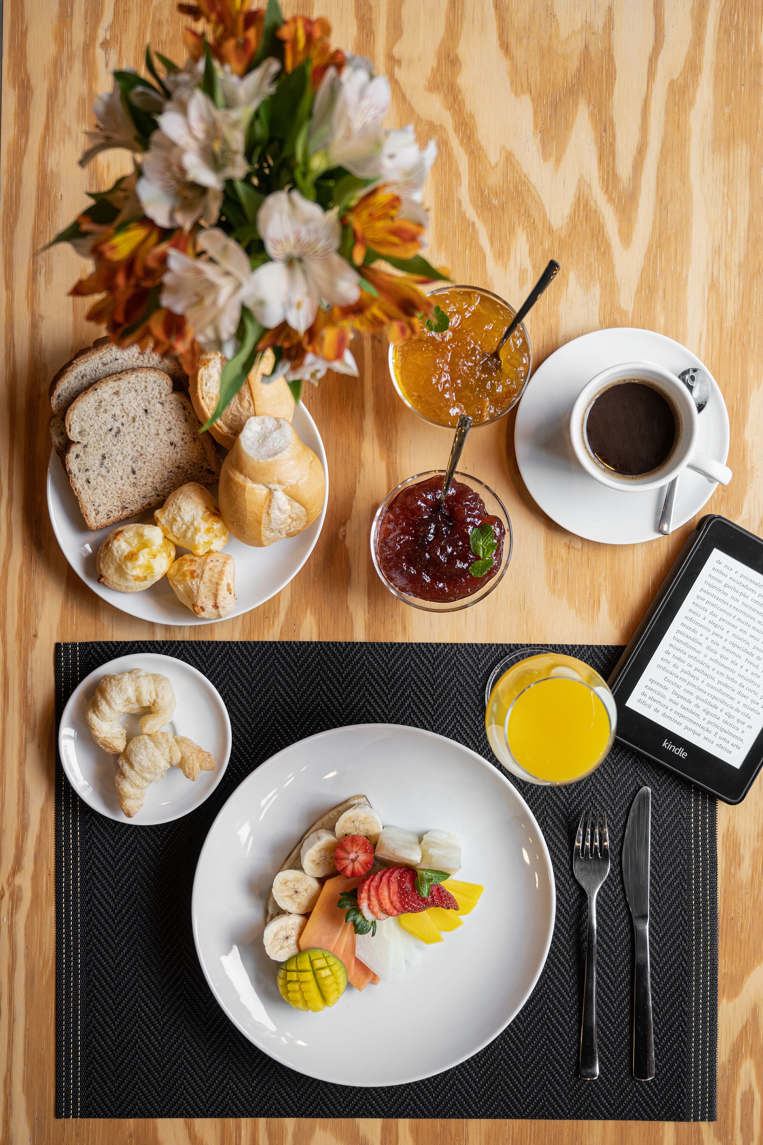 Desayuno con cruasanes, una bandeja de frutas, un vaso de zumo de naranja, una taza de café y una tableta mostrando texto.