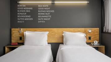 Quarto de hotel com duas camas, cabeceira de madeira, luminária na parede e mesa de cabeceira com porta-retratos.
