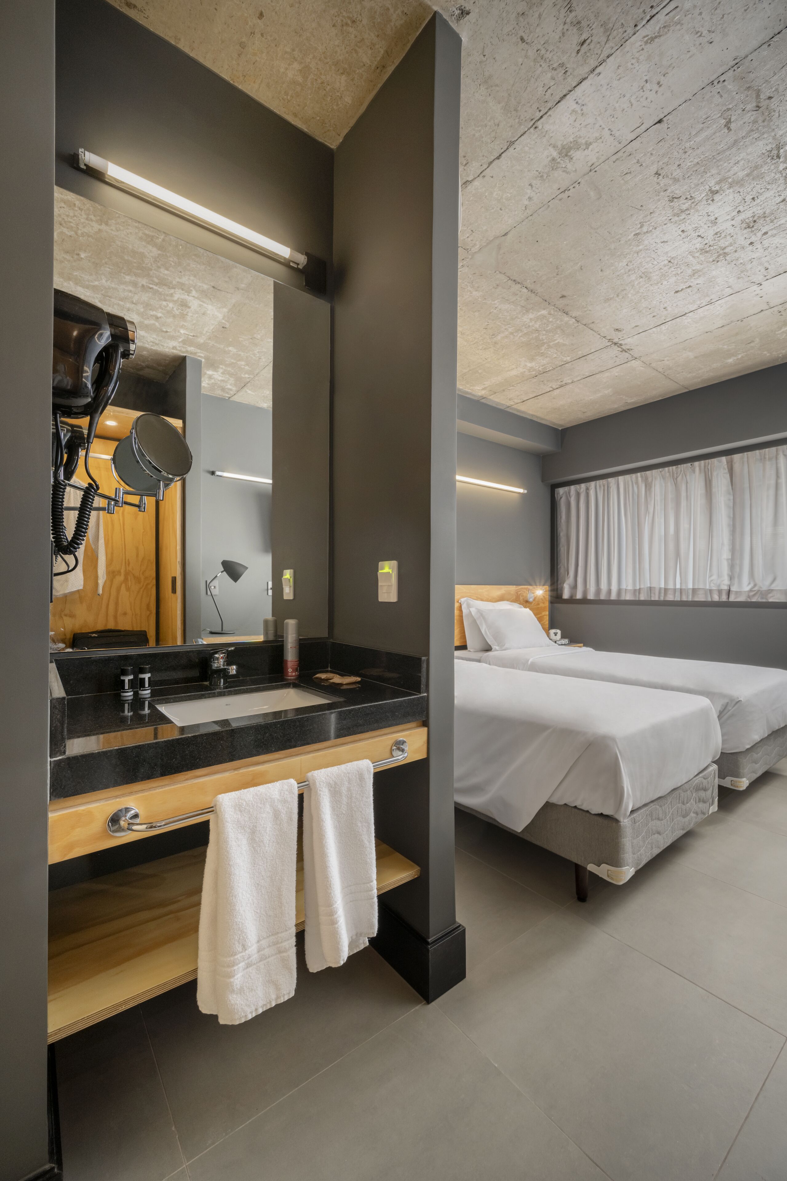 Quarto de hotel moderno com um espelho grande, uma pia, duas camas e teto com vigas aparentes.