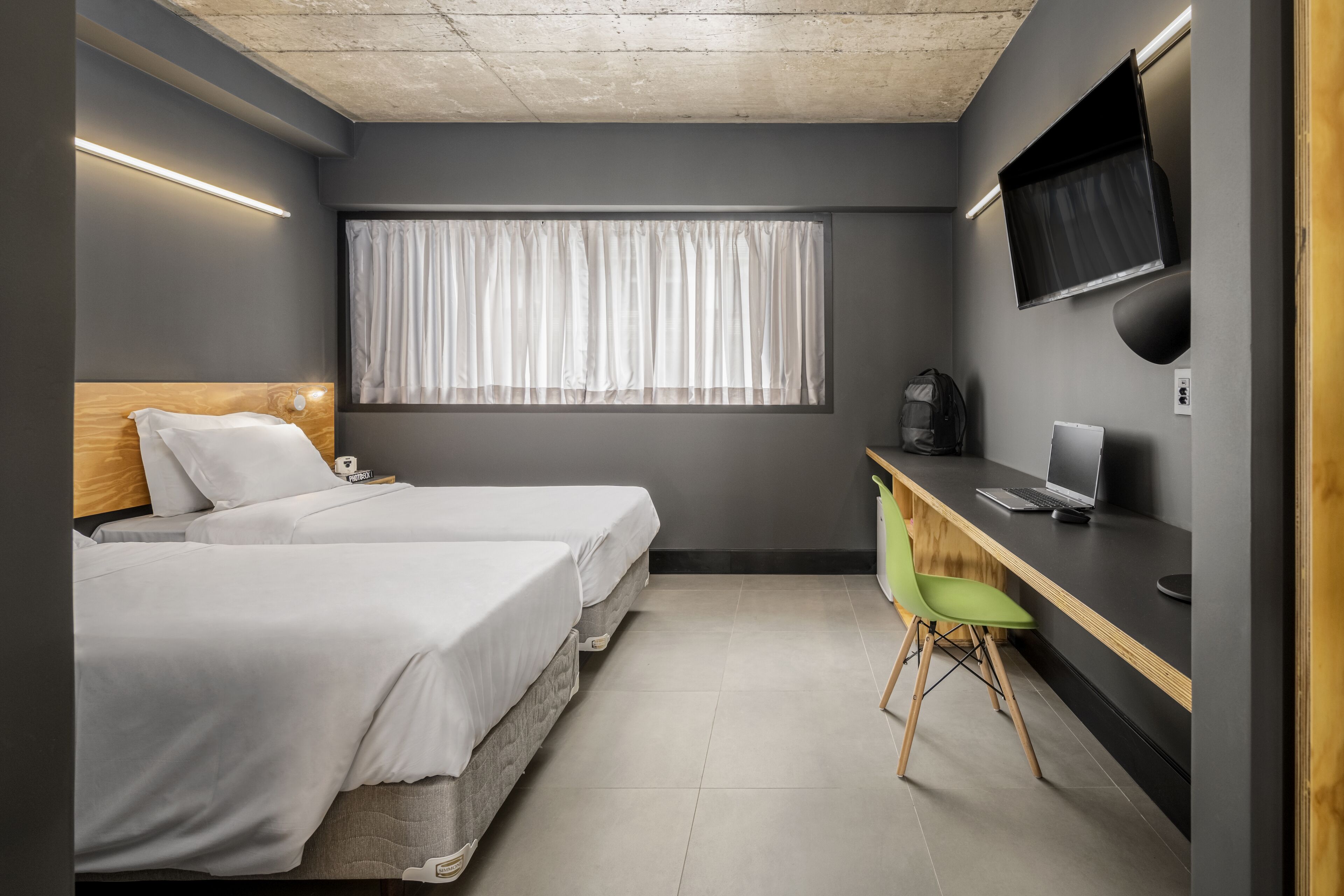 A compact hotel room with two beds, a desk with a laptop, a chair, a TV mounted on the wall, and a window with curtains.
