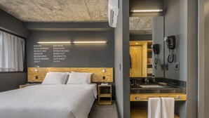 Una habitación de hotel moderna con una cama grande, mesitas de noche, un cabecero de madera, un escritorio integrado con un espejo y un baño con lavamanos y toallero.