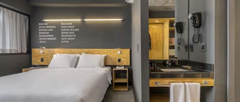 Una habitación de hotel moderna con una cama grande, mesitas de noche, un cabecero de madera, un escritorio integrado con un espejo y un baño con lavamanos y toallero.