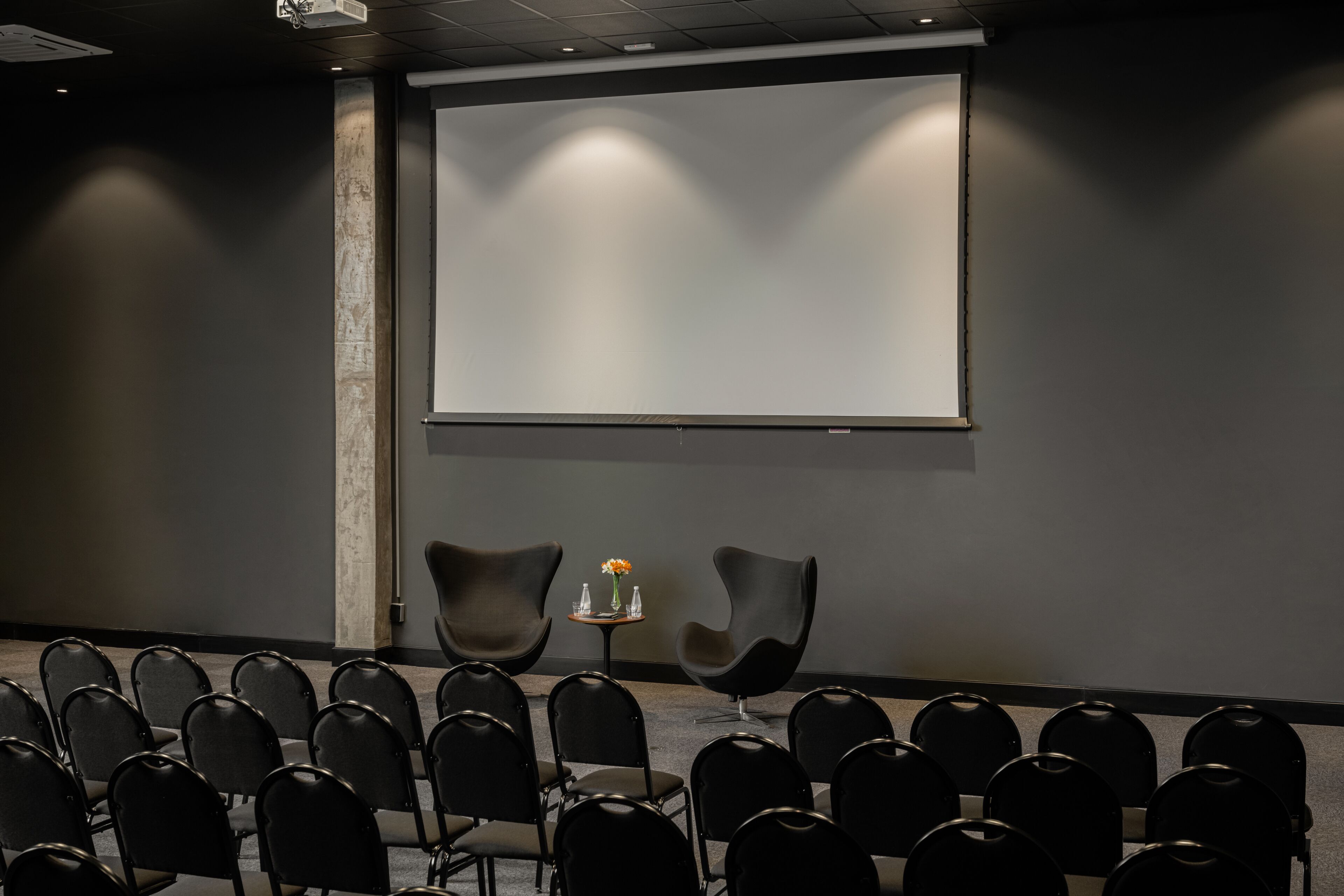 Uma sala de conferências com fileiras de cadeiras pretas, uma tela de projetor e duas cadeiras modernas com uma pequena mesa e um vaso de flores.