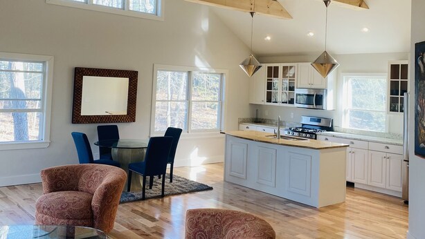 A spacious living area with a wooden ceiling, a dining table with chairs, a kitchen with white cabinets, and a large window. - Reset and Relax in Luxury Cottage (Rhinebeck)