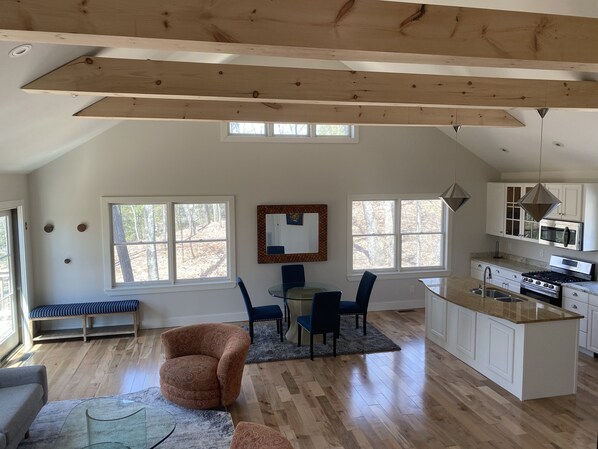 A spacious living area with a high ceiling, wooden beams, a dining table with chairs, a kitchen with white cabinets, and large windows offering views of trees. - Reset and Relax in Luxury Cottage (Rhinebeck)