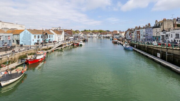 A harbor with boats docked along the waterfront, buildings lining the shore, and a clear sky above. - The Chocolate Box (Weymouth)