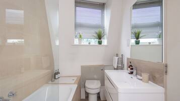 A modern bathroom with a bathtub, toilet, and sink. There are two windows with blinds and potted plants.