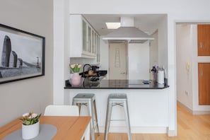 A modern kitchen with white cabinets, a black countertop, and a stainless steel range hood. There are two bar stools and a dining table with a vase of flowers.
