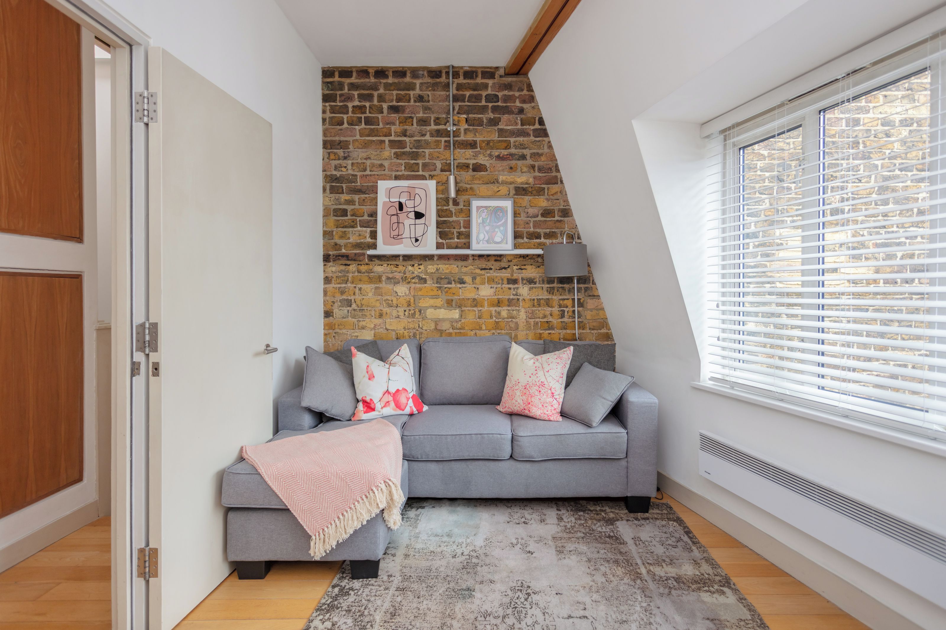 A modern living room with a grey sofa, a patterned rug, a brick wall, and a window with blinds.