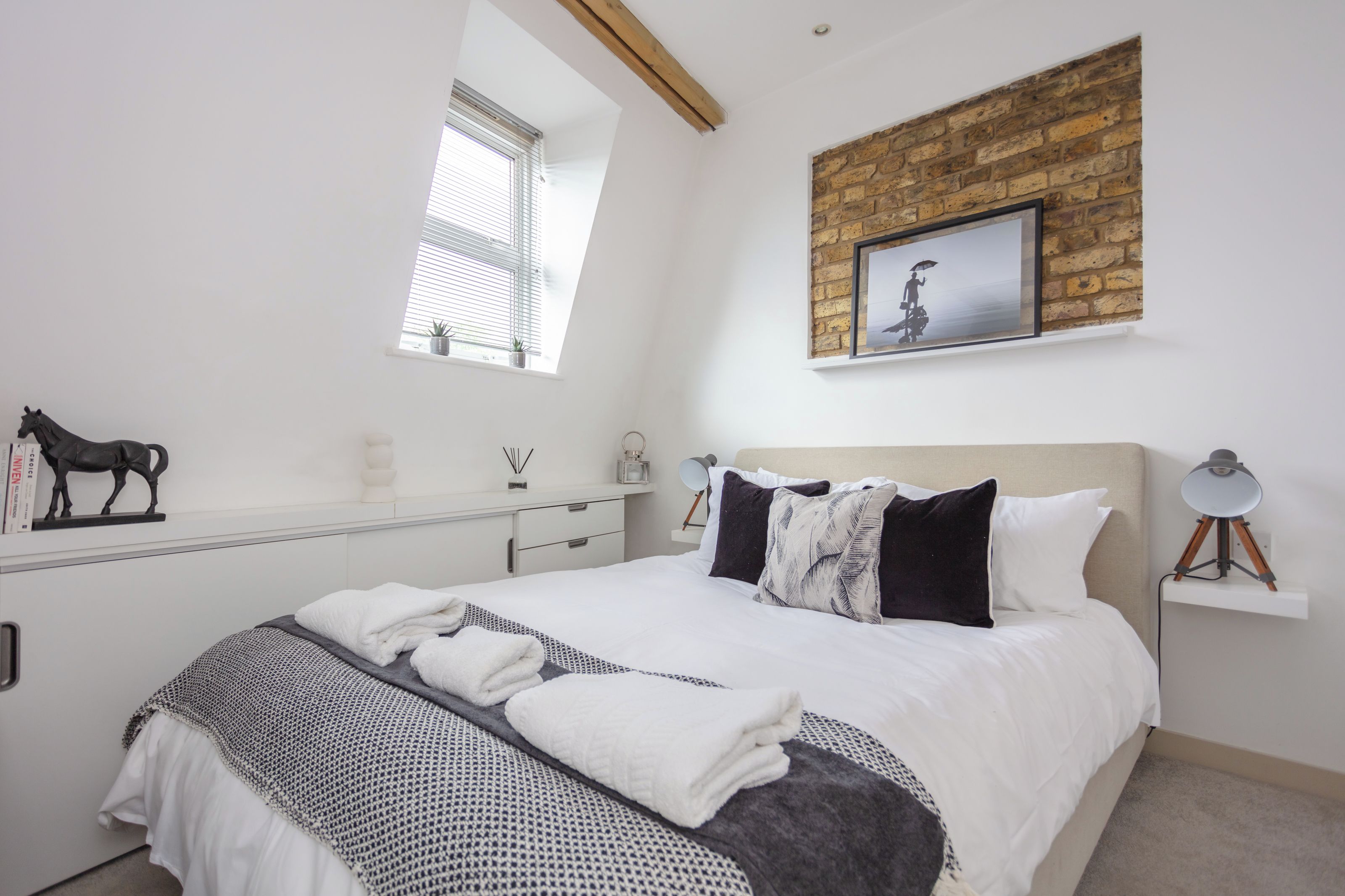 Een slaapkamer met een bed, nachtkastje, lamp, raam en een bakstenen muur.