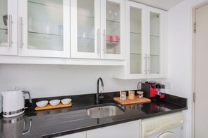 A kitchen with white cabinets, a black countertop, a stainless steel sink, a coffee maker, and a red and black coffee machine. - Stylish 1BD by Northcote Road and Clapham Junction (London)