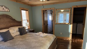 Une chambre avec un grand lit, une tête de lit en bois et une fenêtre avec des stores. Une porte donne accès à une salle de bain et à un coin cuisine.