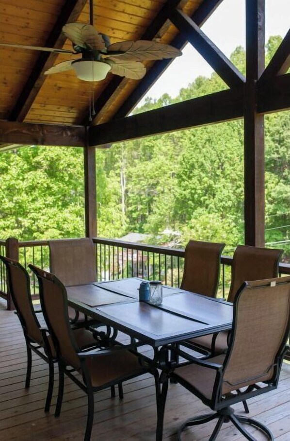 A covered outdoor dining area with a table and chairs, a ceiling fan, and a view of a forested area.
