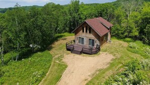 Une cabane en bois avec un toit rouge, entourée d’une végétation luxuriante et avec des collines en arrière-plan.