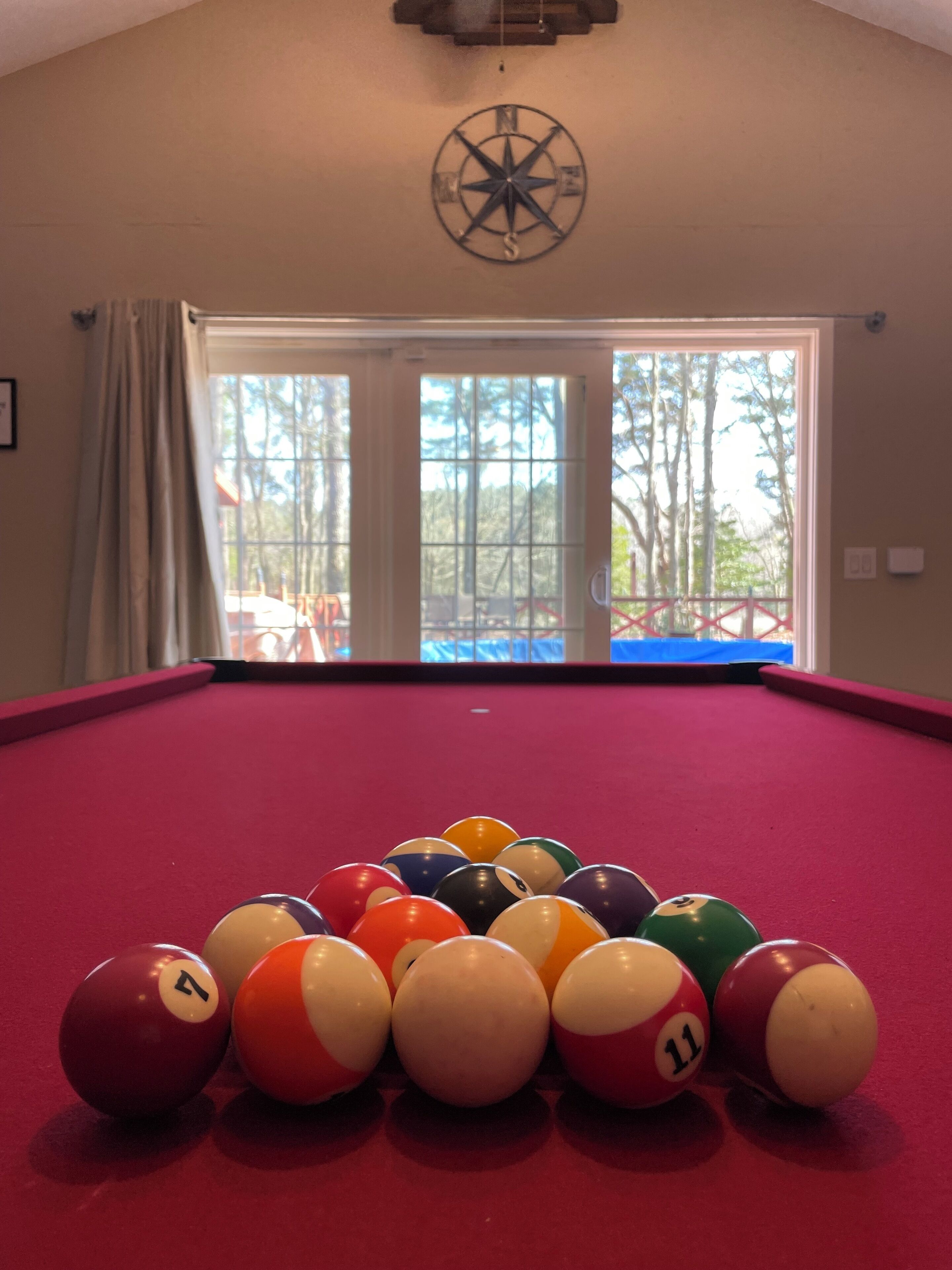 Une table de billard avec les billes disposées en forme de triangle, une décoration murale représentant une boussole et une vue sur les arbres à travers une grande fenêtre.