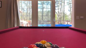 Une table de billard avec les billes disposées en forme de triangle, une décoration murale représentant une boussole et une vue sur les arbres à travers une grande fenêtre.