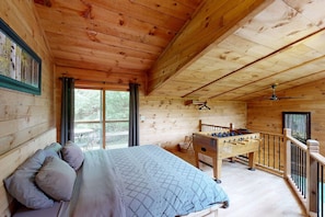 Une pièce chaleureuse, aménagée dans un chalet en bois, avec un lit, une table de baby-foot et un balcon offrant une vue sur les arbres.