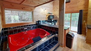A cabin interior with a red jacuzzi, a kitchen area, and a large window overlooking a forest. - The Hideaway (Norwalk)