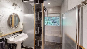 A modern bathroom with a pedestal sink, a shower with a glass door, and a view of a forest through the shower. - The Hideaway (Norwalk)