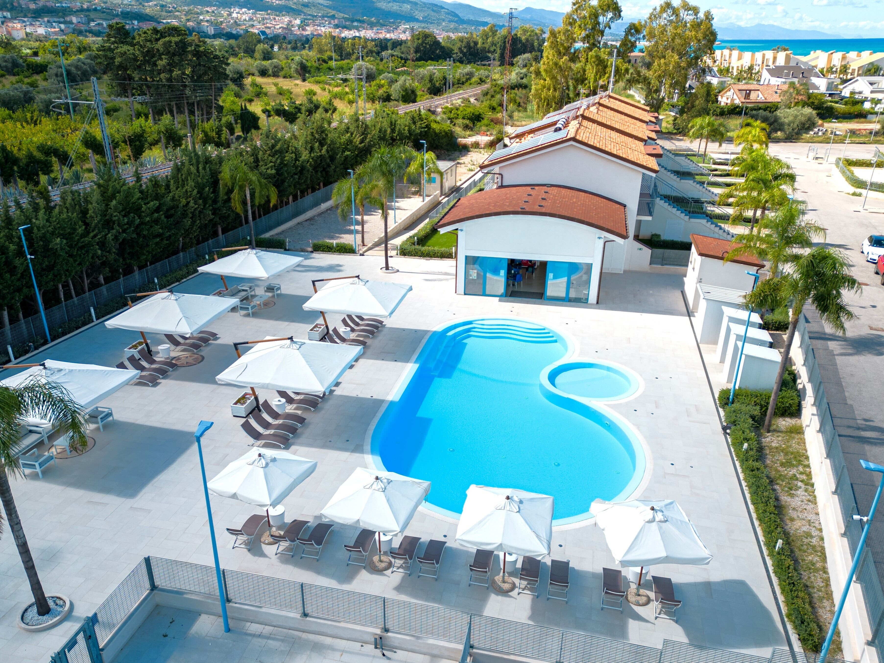 Zona de piscina con sillas de descanso y sombrillas, rodeada por un edificio de techo rojo.