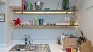 Una cocina con un lavamanos, una rejilla metálica y estantes superiores que sostienen libros, utensilios de cocina y un juguete rojo.