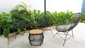 Drei schwarze Stühle aus Drahtgestell und ein runder Holztisch auf einer Beton-Terrasse mit Grünpflanzen.