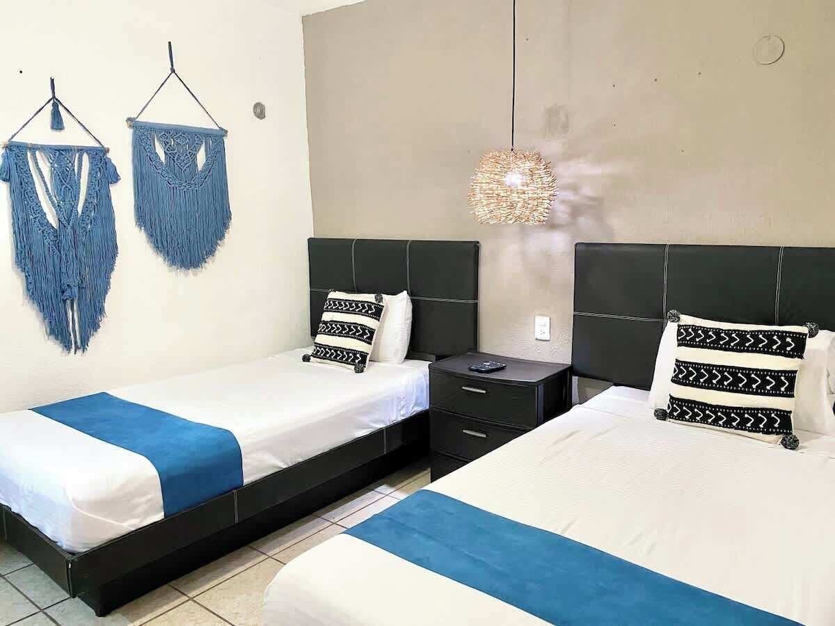 A hotel room with two beds, a nightstand, a hanging light fixture, and a wall-mounted macrame decoration.