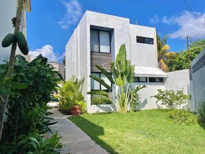 A modern two-story building with a flat roof, surrounded by a well-maintained garden with various plants and a paved pathway.