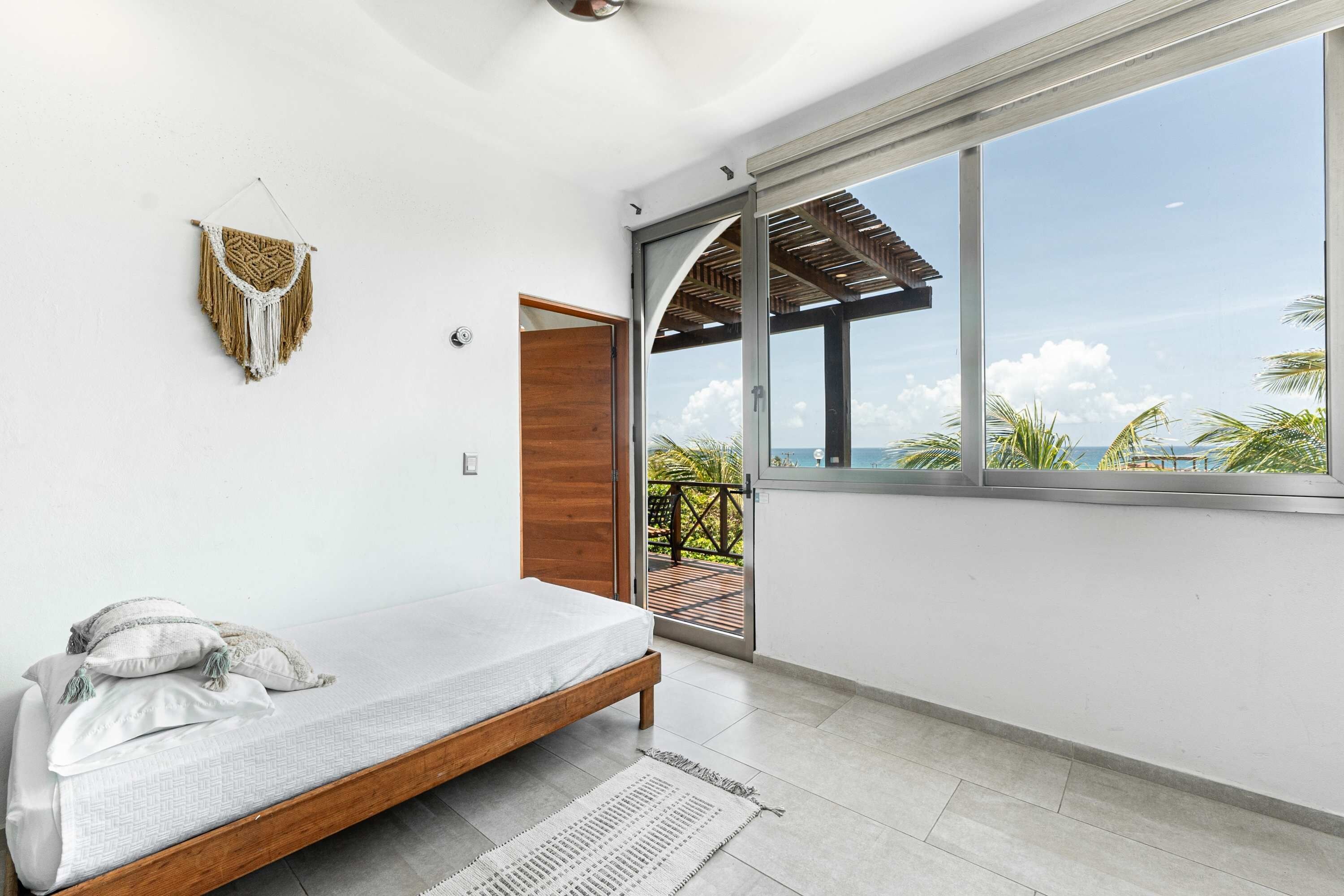 A modern bedroom with a bed, a wall-mounted macrame piece, and a large window with a view of the ocean.