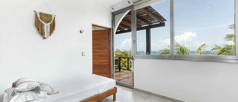 A modern bedroom with a bed, a wall-mounted macrame piece, and a large window with a view of the ocean.
