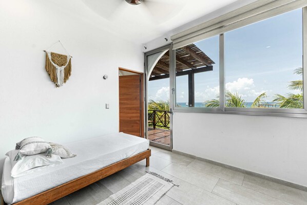 A modern bedroom with a bed, a wall-mounted macrame piece, and a large window with a view of the ocean.