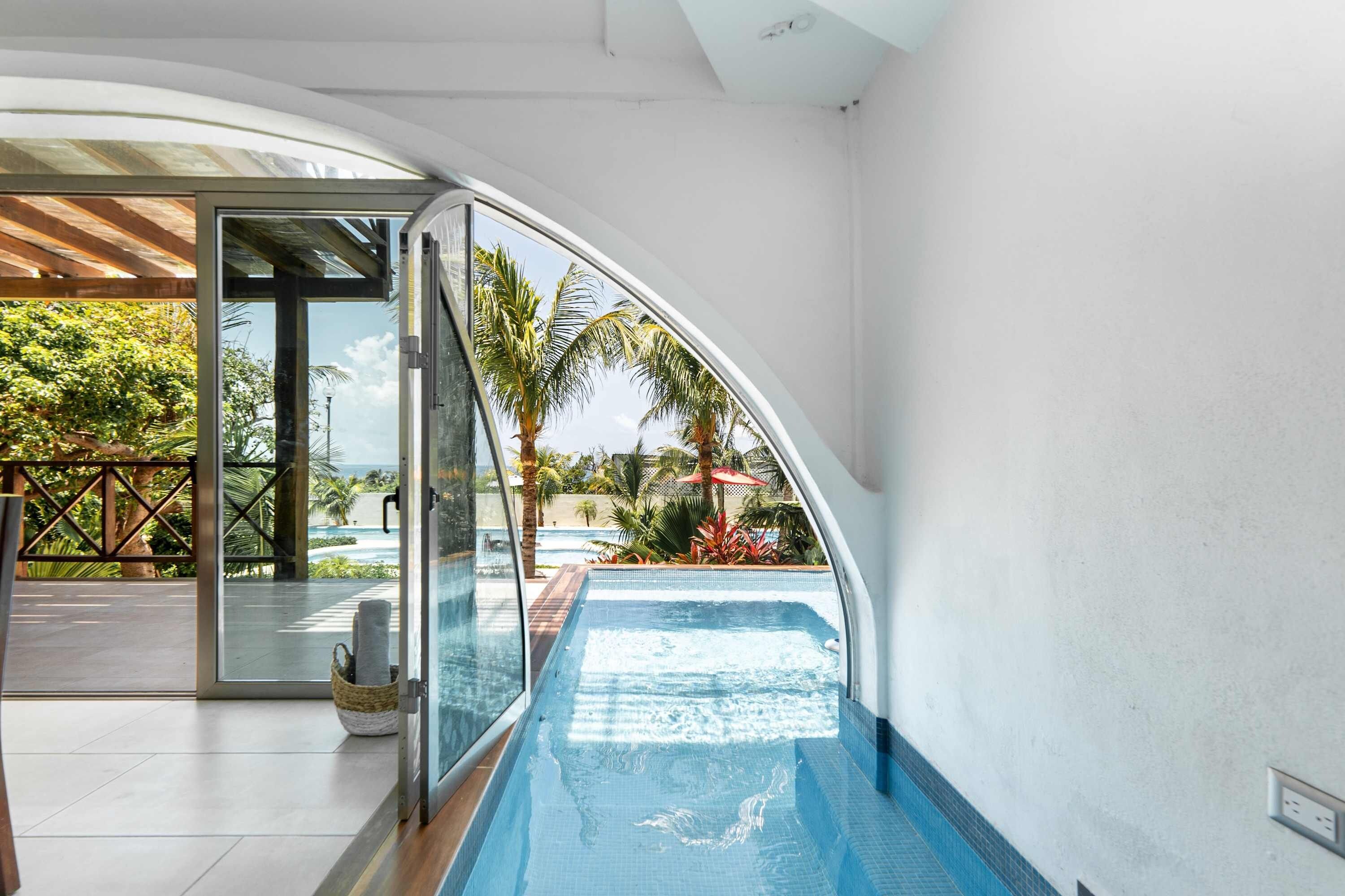 A modern indoor swimming pool with a clear view of the outdoors, including a poolside area and palm trees.