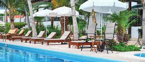Un área junto a la piscina con sillas de descanso y sombrillas, rodeada de palmeras.