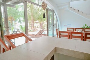 A modern interior with a large table and chairs, a hanging light fixture, and a view of palm trees through large windows.