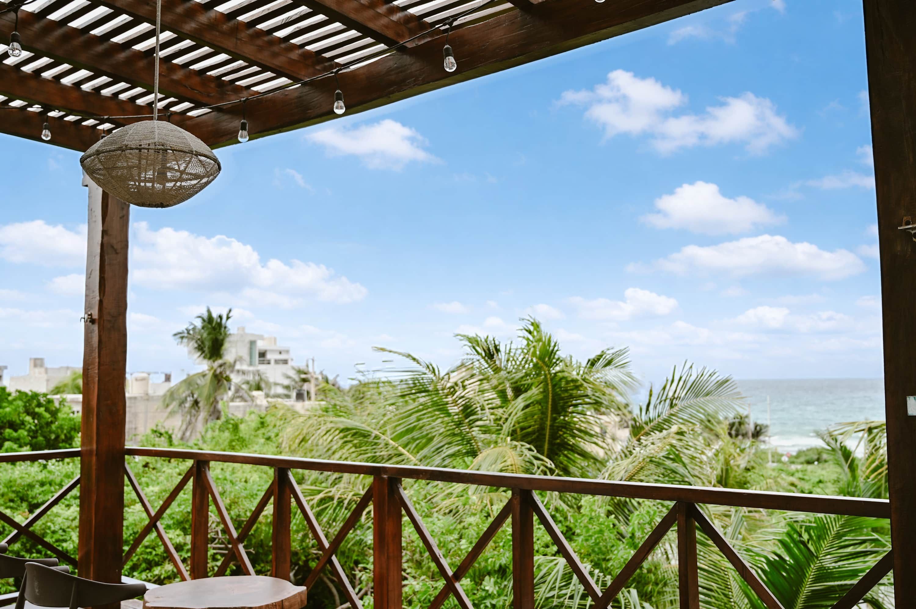 A wooden pergola with a hanging light fixture, a view of the ocean, and palm trees.