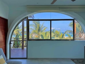 A room with a large arched window overlooking a deck with palm trees and a view of the ocean.