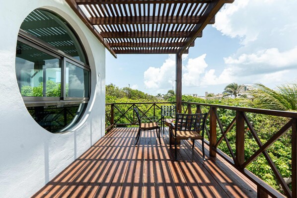 A wooden deck with a pergola, a round window, and a view of greenery and buildings.