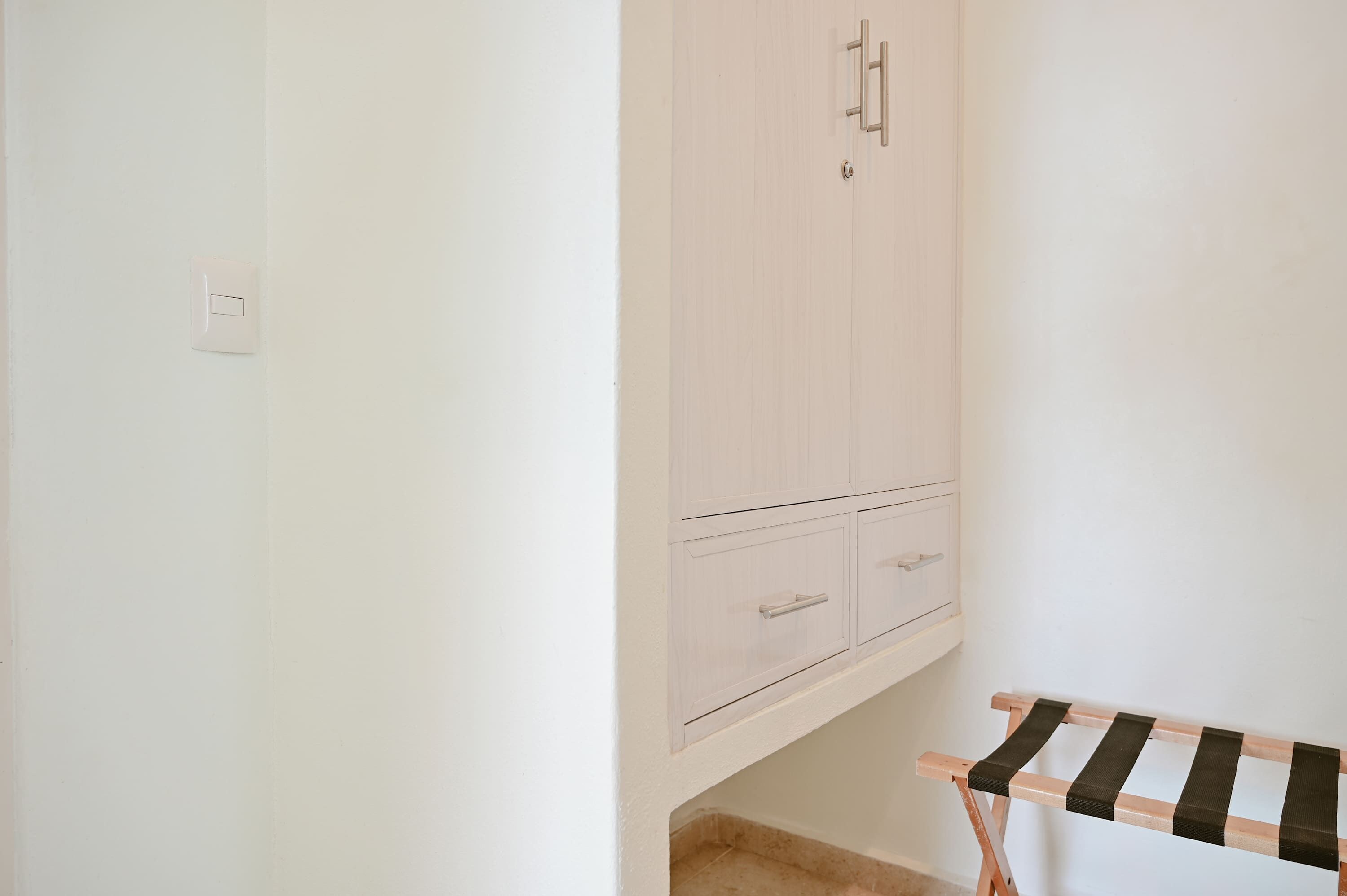 A room with a white wardrobe, a small table with a striped cloth, and a light switch on the wall.