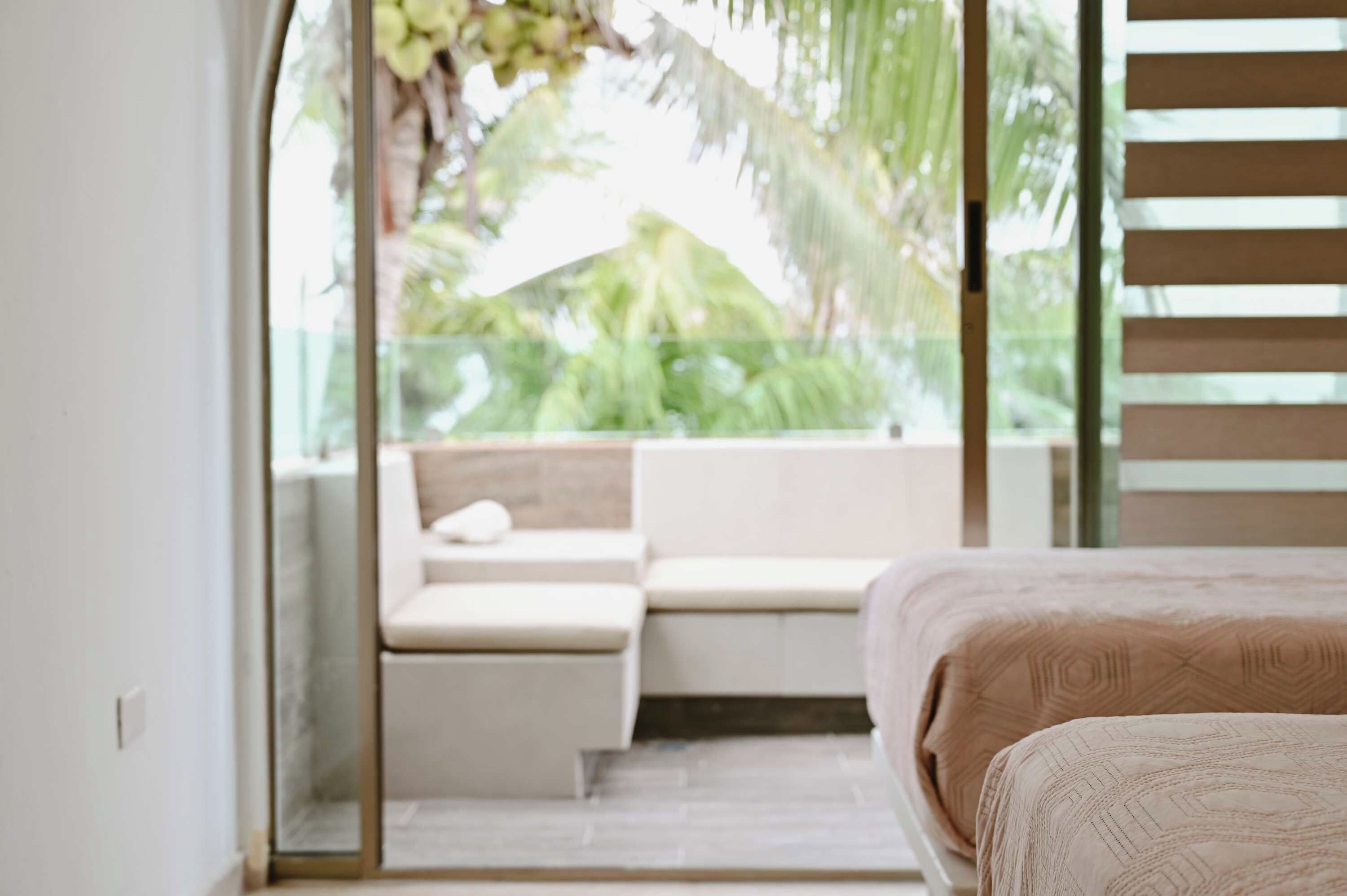 A bedroom with a bed, a seating area with a bench, and a view of greenery through a glass door.