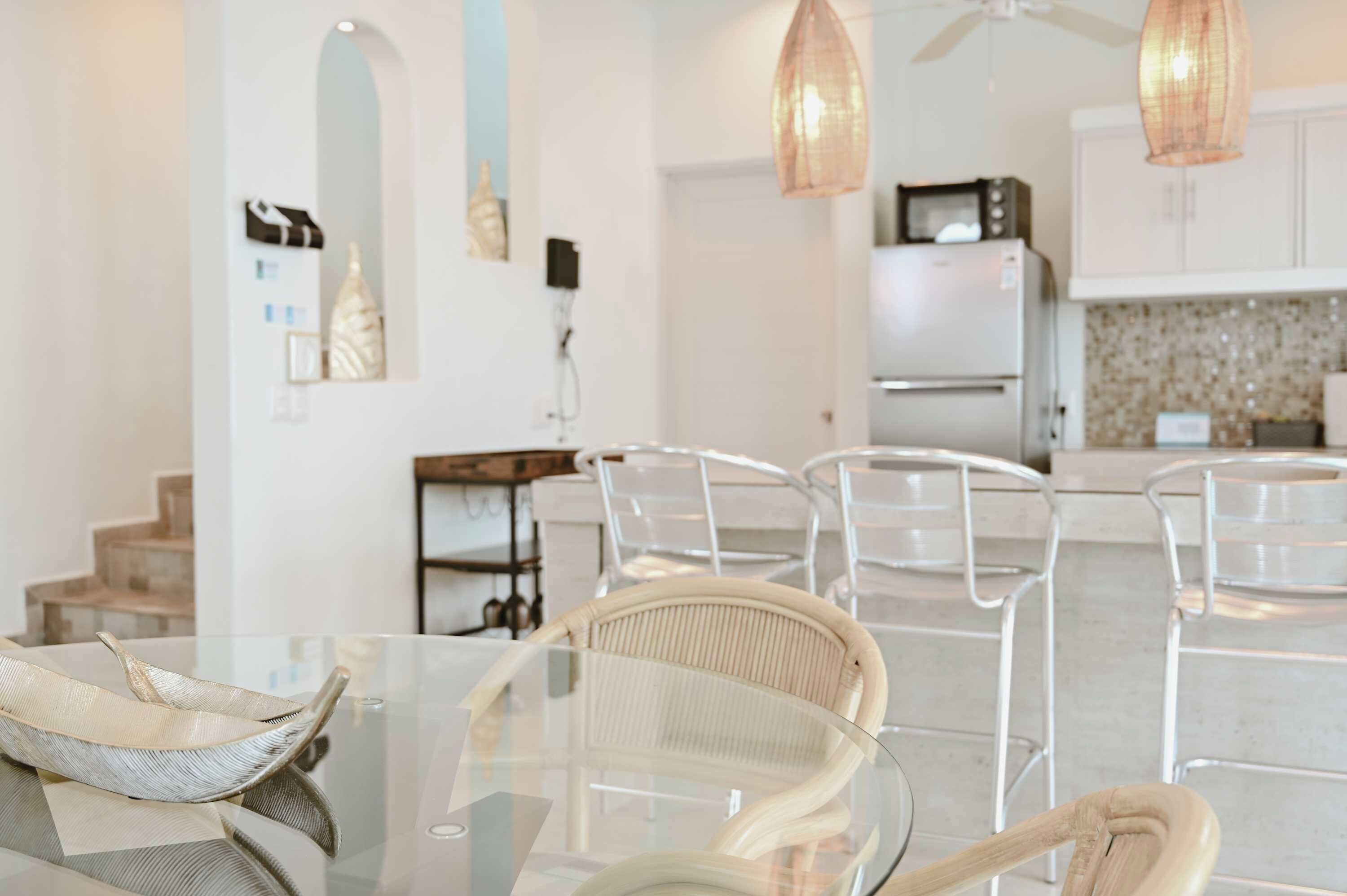 A modern kitchen with white cabinets, a microwave, and a refrigerator. There are clear chairs around a glass dining table.