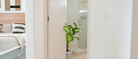 Una habitación de hotel con cama, mesita de noche, planta en maceta y zona de ducha.