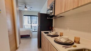 Eine kleine Küche mit Mikrowelle, Herd und Spüle. Im angrenzenden Raum ist ein Bett zu sehen, und ein Balkon ist durch eine offene Tür erreichbar.