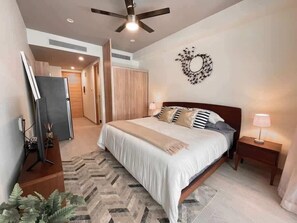 A bedroom with a bed, bedside tables, a ceiling fan, a wall decoration, and a hallway. - Playa Studio | Rooftop Pool, Steps to 5th Ave (Playa del Carmen)