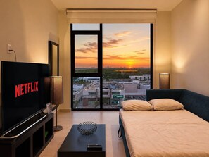 A modern hotel room with a large TV displaying 'NETFLIX,' a bed, a coffee table with a remote, and a view of a sunset through floor-to-ceiling windows.