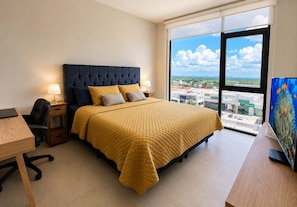 A bedroom with a bed, a desk, a chair, a TV, and a large window with a city view.