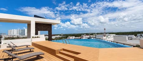 A rooftop pool with wooden deck and lounge chairs, a covered seating area, and a cityscape view.