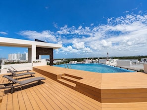 A rooftop pool with wooden deck and lounge chairs, a covered seating area, and a cityscape view.